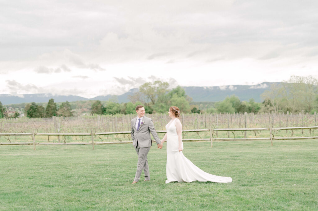 bride and groom walk across lawn with vineyards in the background in VA