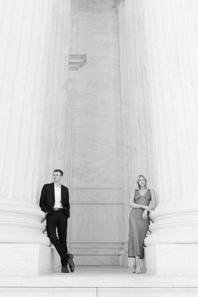 A chic black and white image of a couple leaning against the columns outside of the U.S. Supreme court during their engagement photos.
