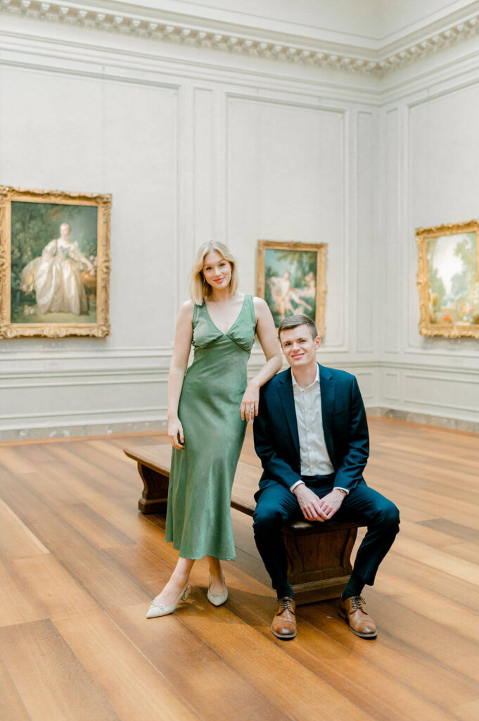 A man and a woman posing together in the National Art Gallery. He is sitting on a bench while she leans onto him, wearing a green satin dress.