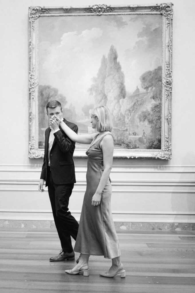 Black and white image of a couple walking through an art gallery as groom kisses his fiance's hand. 