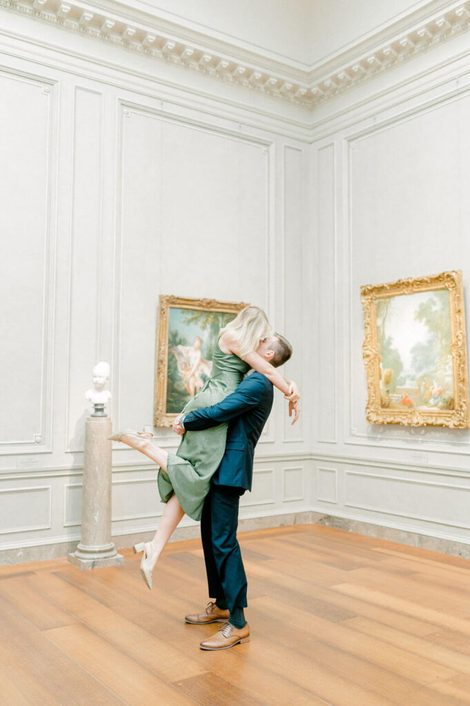 Man lifting his fiance to kiss her in an art gallery.