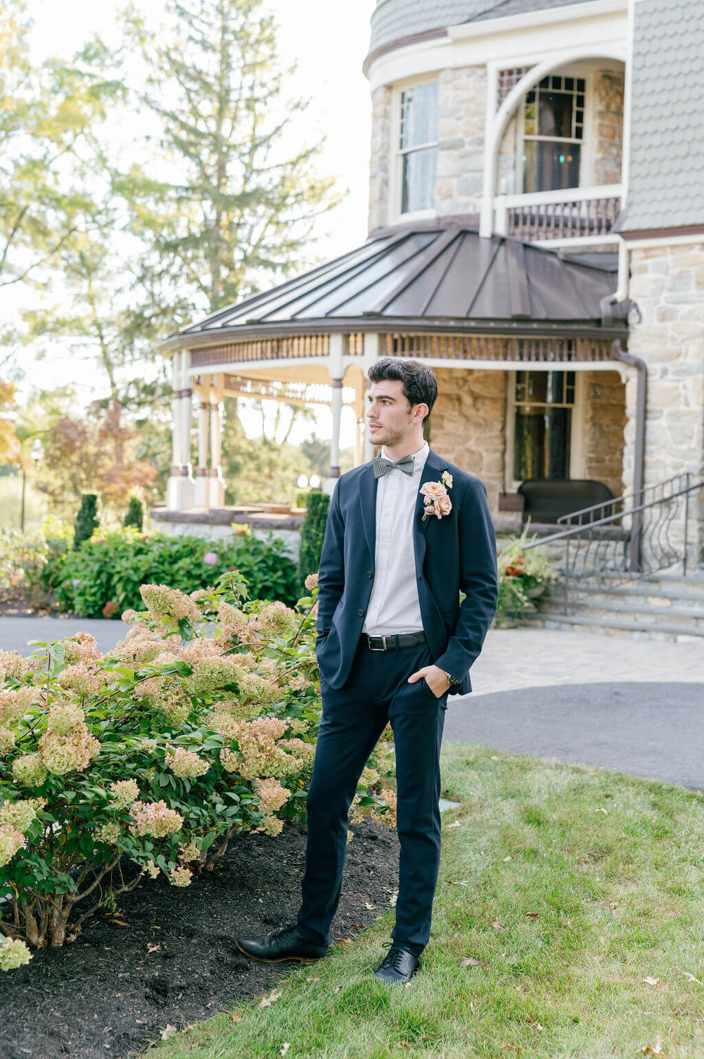 Groom posing outside The Willows at Ashcombe Mansion near blooming hydrangeas and historic stone architecture.