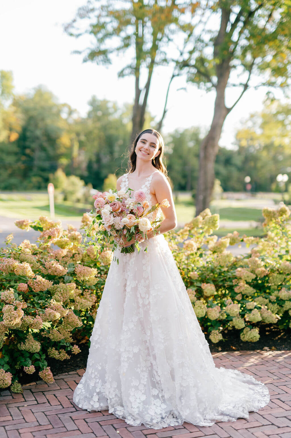 Bride in floral lace gown holding a blush-toned bouquet, smiling beside blooming garden at The Willows at Ashcombe Mansion