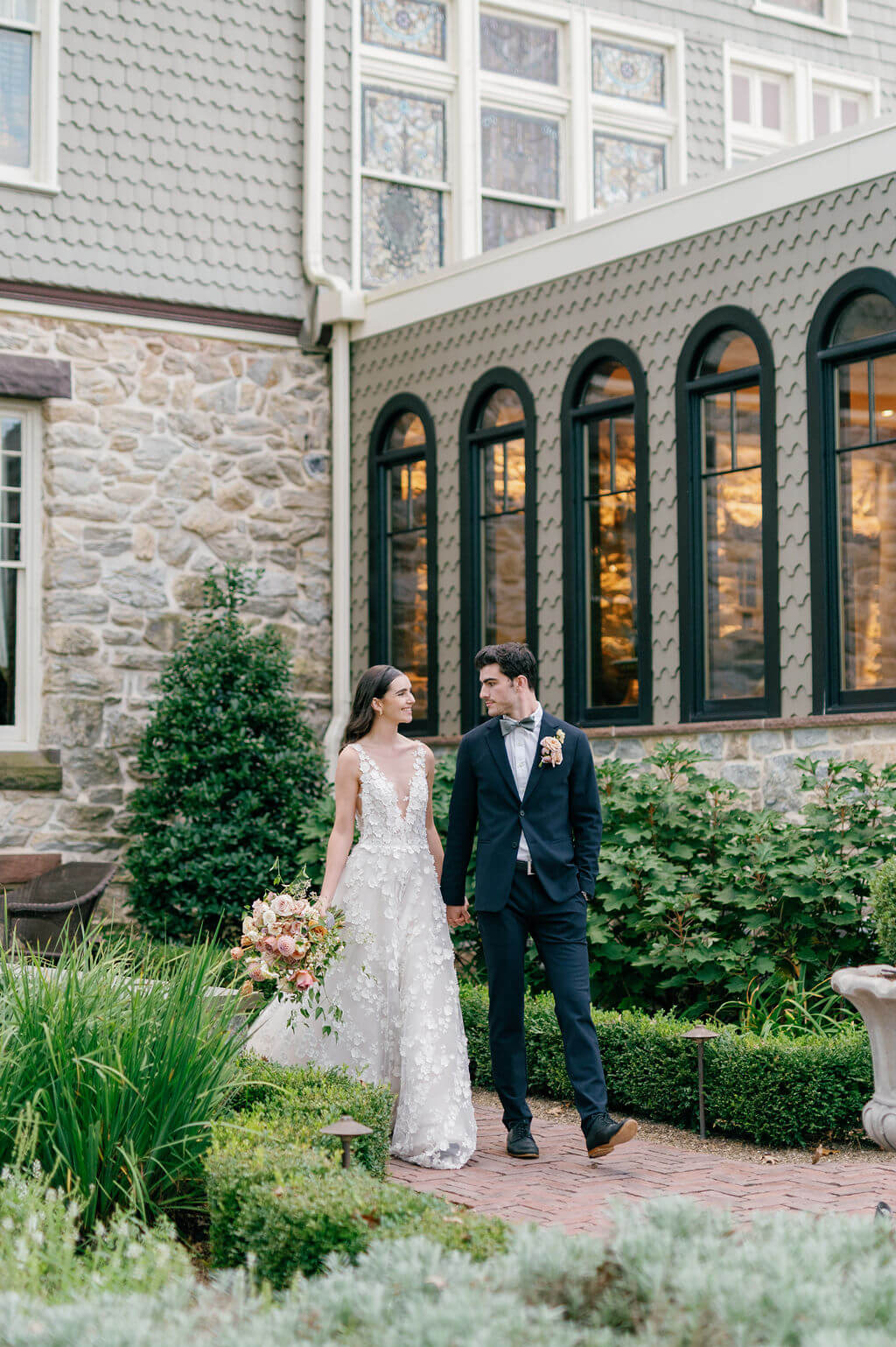 Bride and groom walking hand in hand in the gardens of The Willows at Ashcombe Mansion near the historic stone exterior.