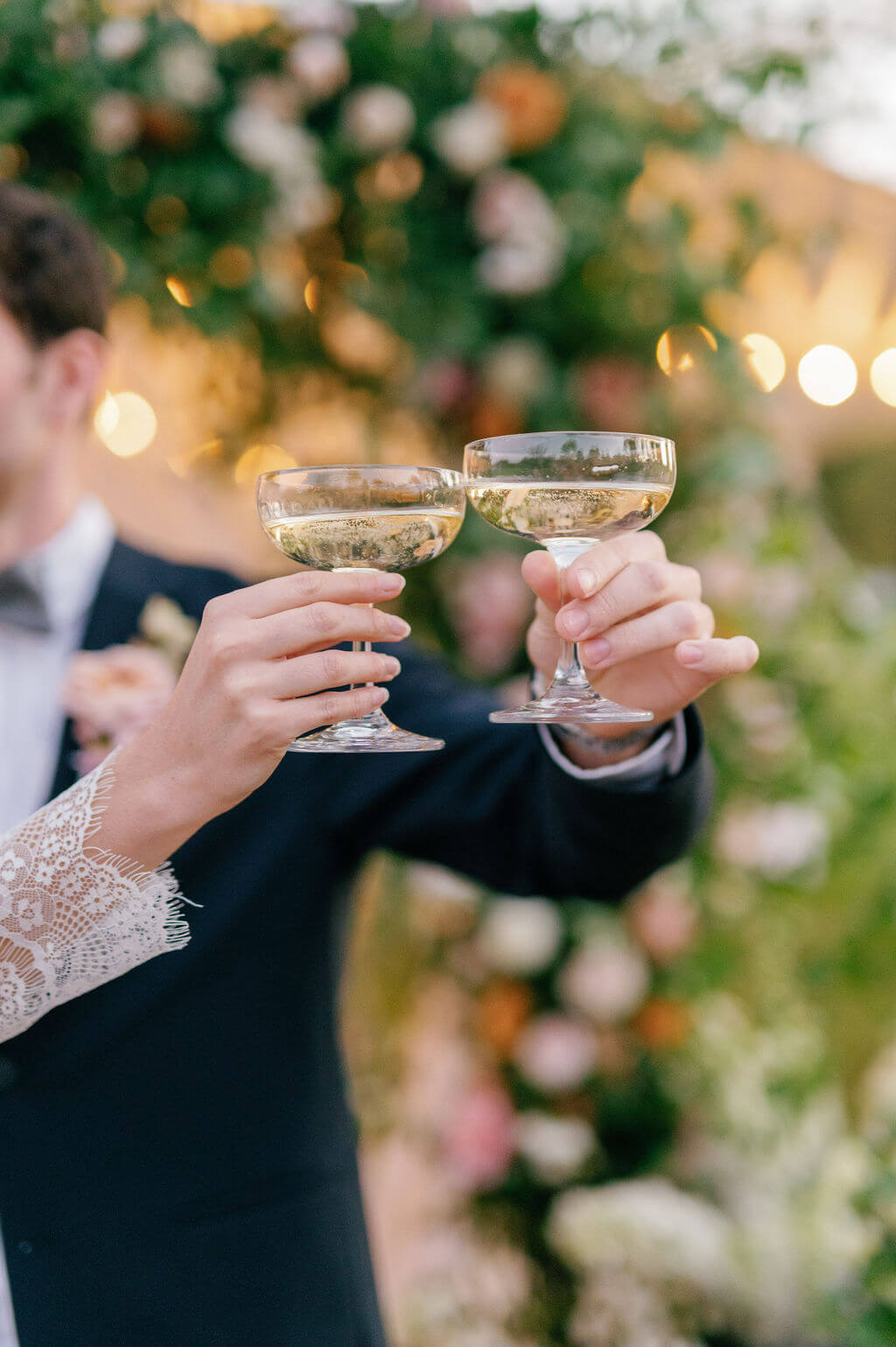 Close-up of bride and groom raising champagne coupes during wedding toast at The Willows at Ashcombe Mansion, with floral backdrop and bokeh lights.