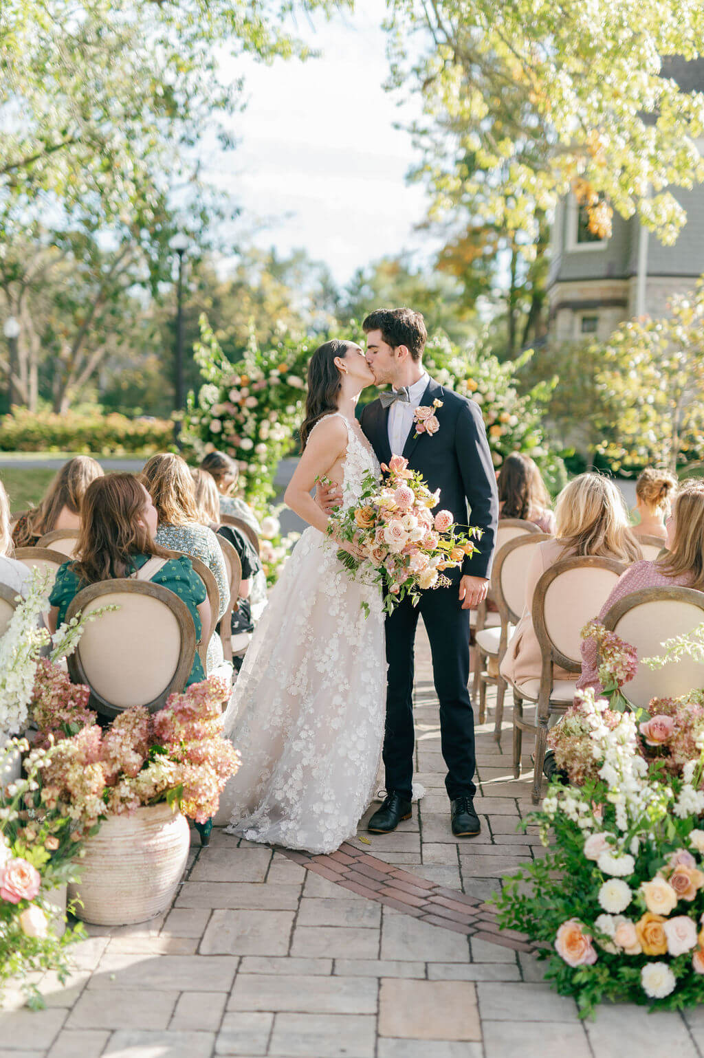 Bride and groom share a kiss during an outdoor ceremony at The Willows at Ashcombe Mansion with floral arrangements and seated guests.