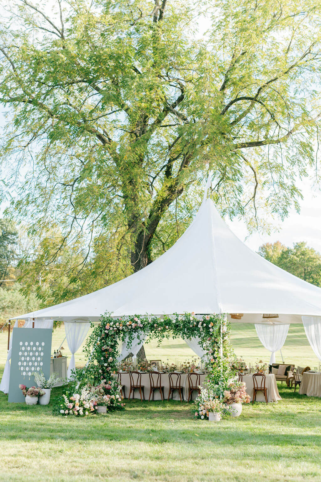 White tented reception at The Willows at Ashcombe Mansion adorned with lush greenery and floral arch, set under a large tree in an open field.