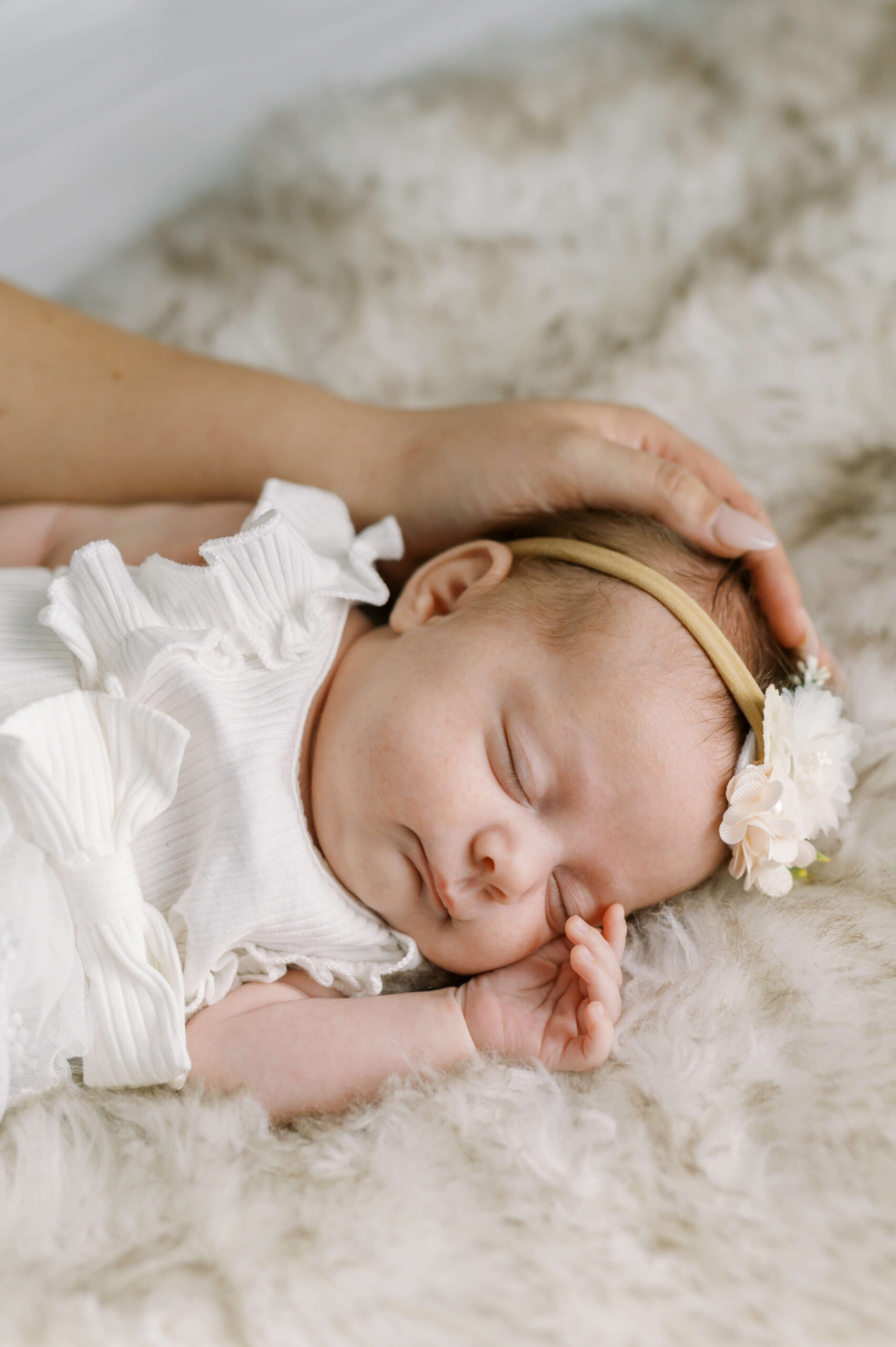 A newborn baby sleeping on a fur blanket in a white dress with matching flower headband