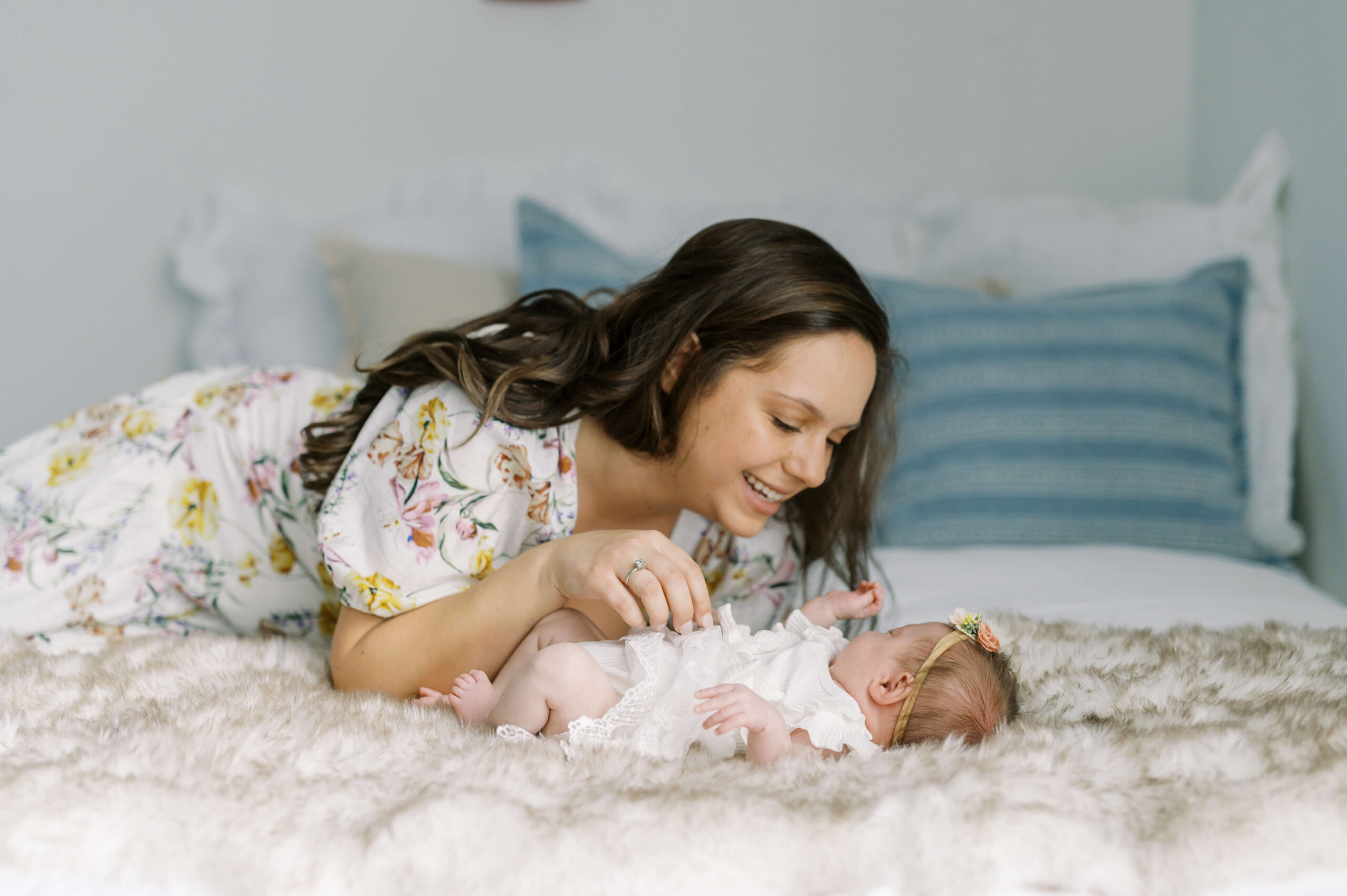 A happy new mom in a flower print dress plays with her newborn on a bed in a white dress after visiting baby boutiques in richmond va