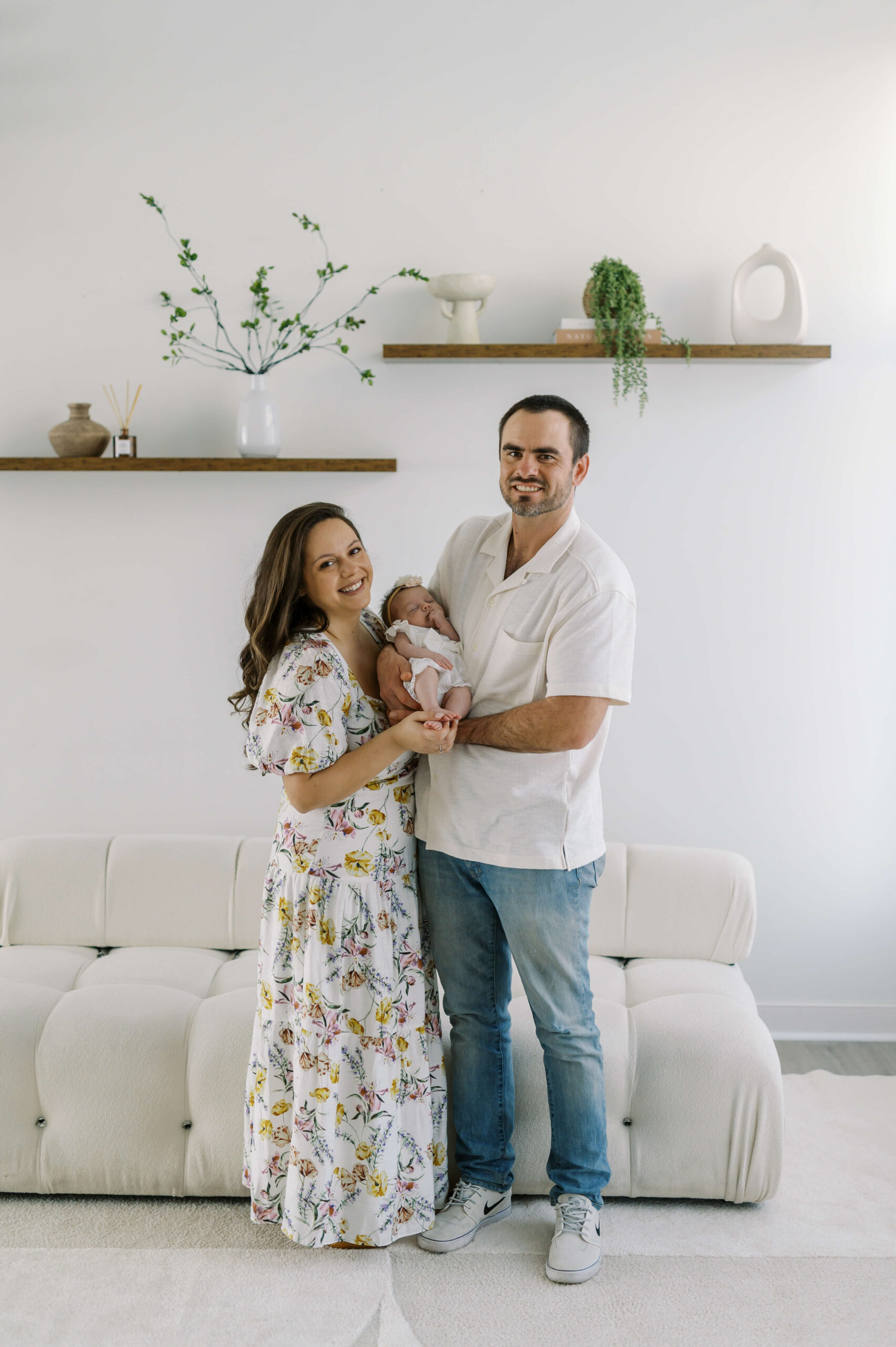 Happy new mom and dad stand in a modern living room holding their sleeping newborn daughter between them after visiting baby boutiques in richmond va