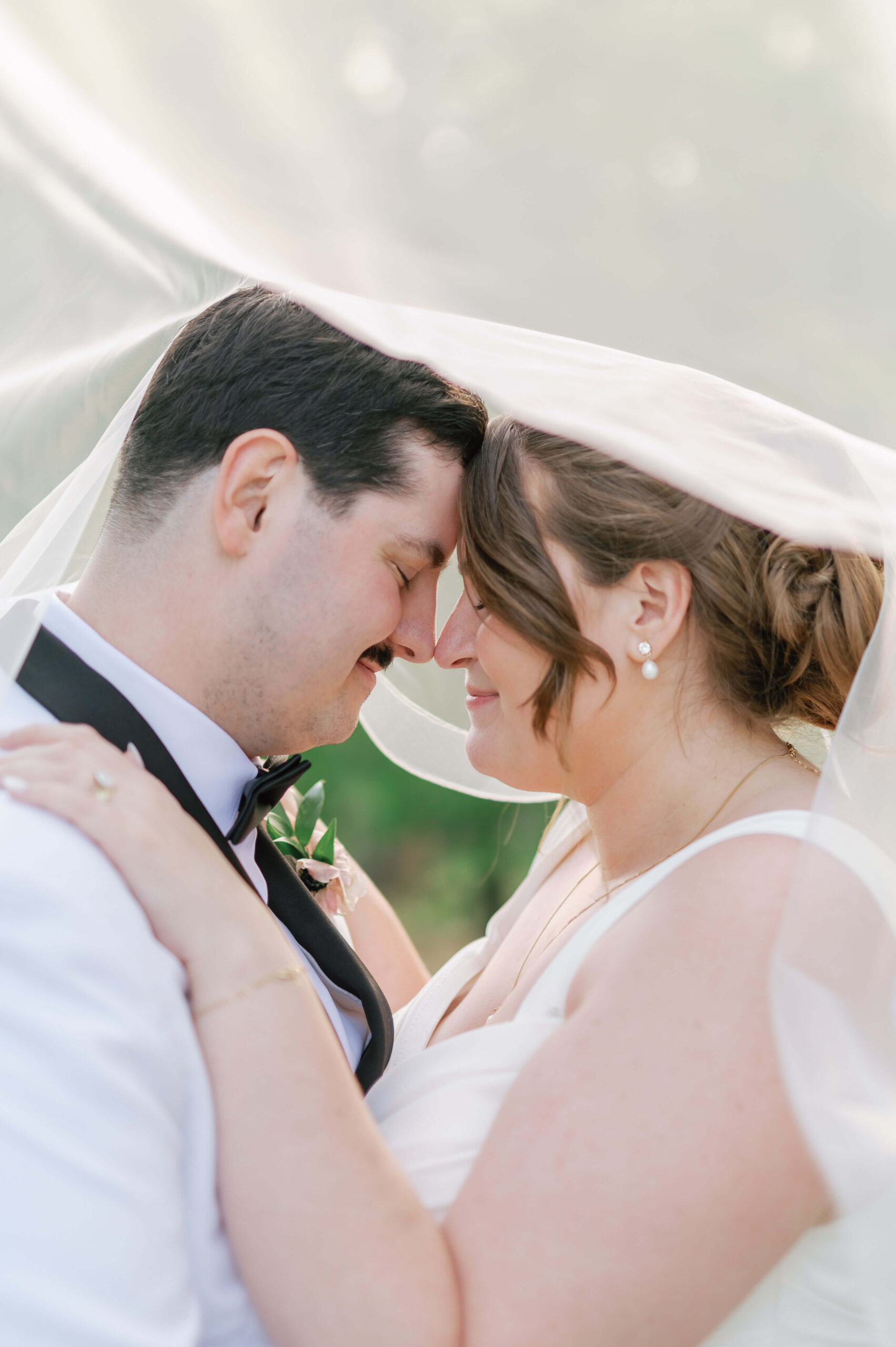 A groom in a black and white tux smiles while touching foreheads with his bride under the veil