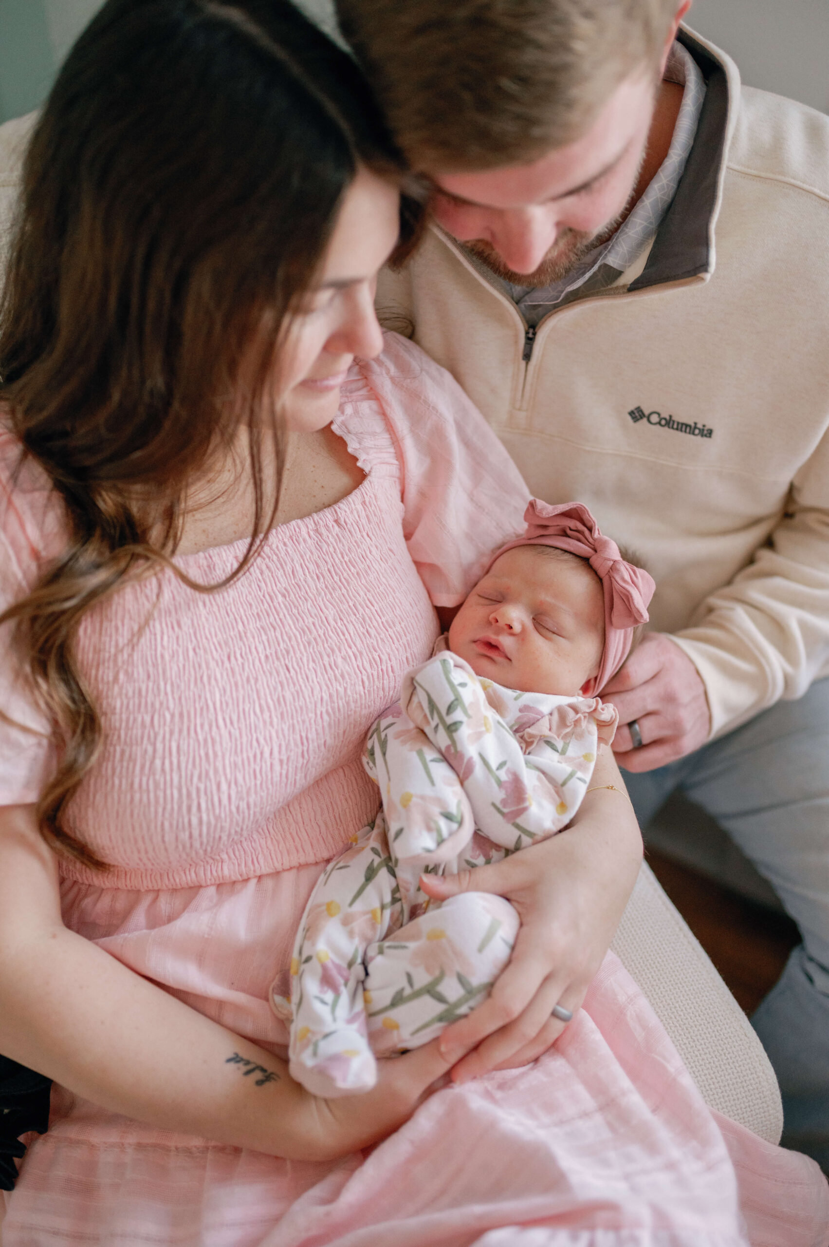 A newborn baby girl in. a flower print swaddle sleeps in mom's arms in a pink dress with dad snuggling in