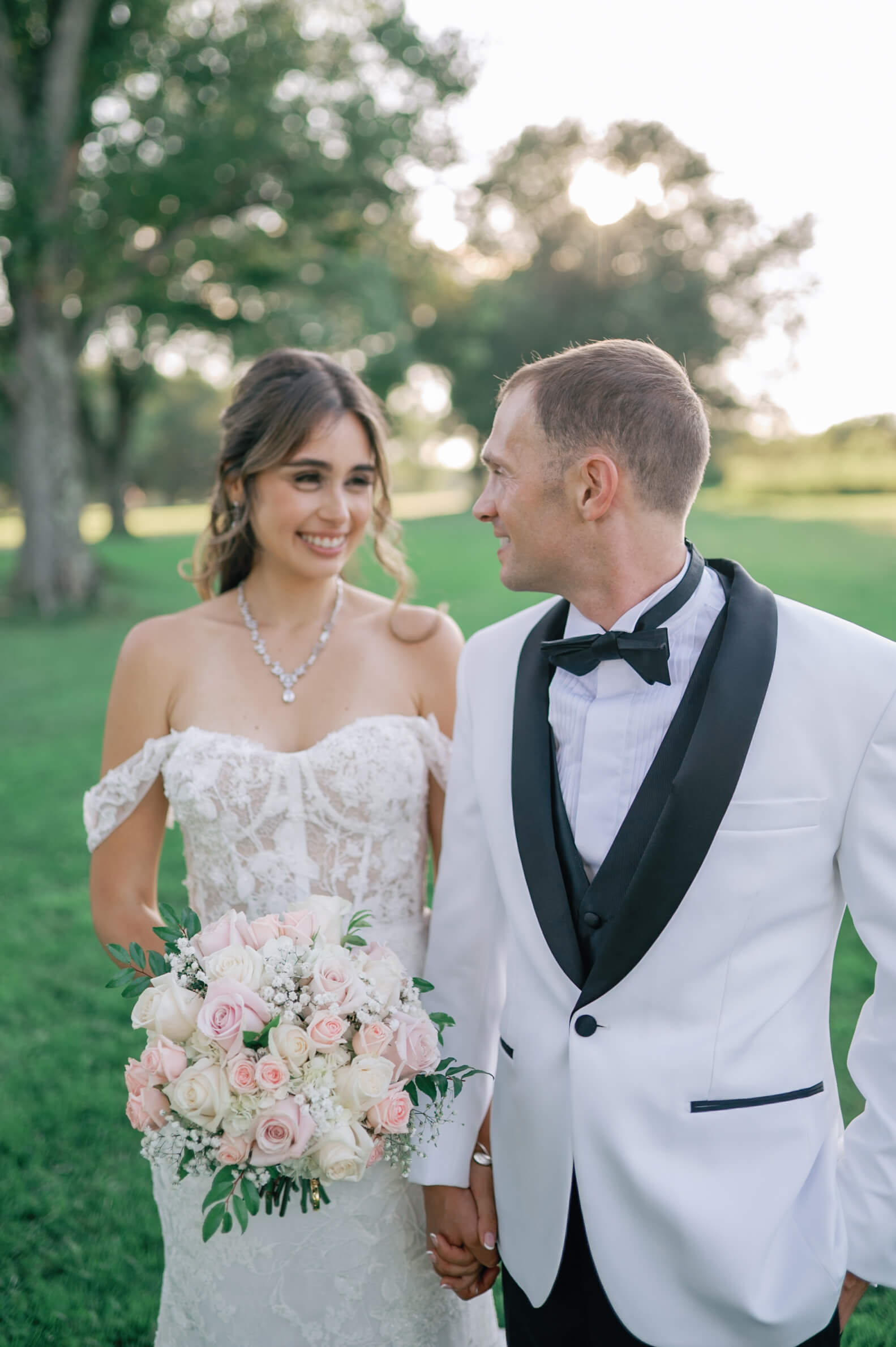 A groom looks back smiling over his shoulder to his happy bride in a lawn holding hands at sunset