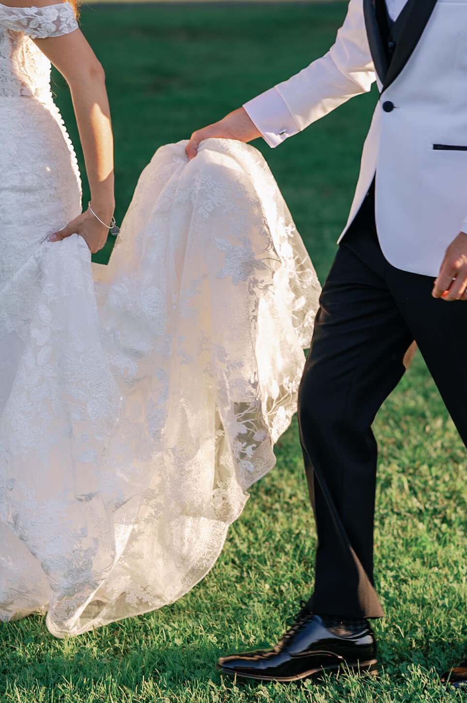 Details of a groom in black and white tux carrying his bride's train in a lawn during their pippin hill farm wedding