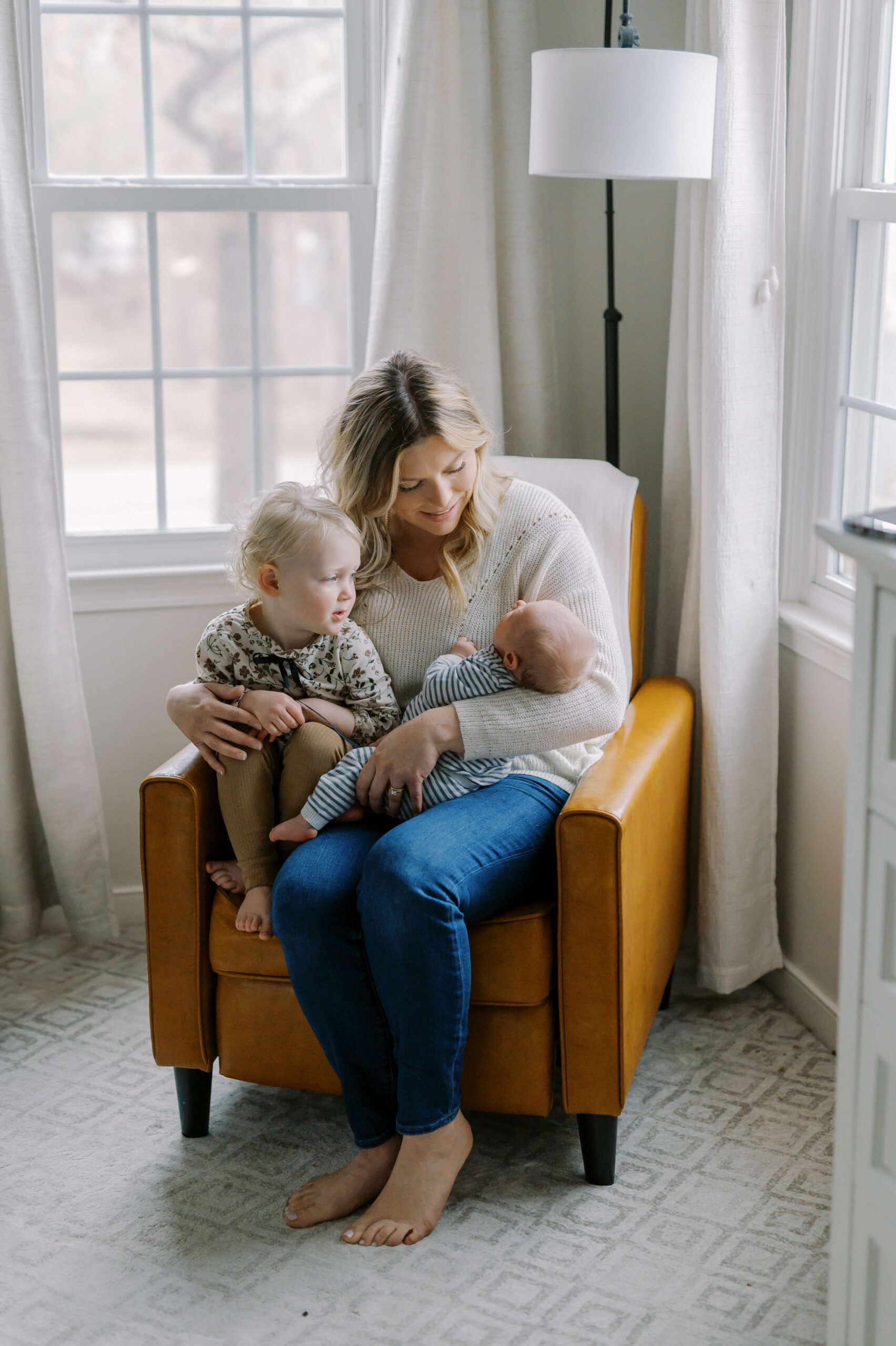 A smiling mother sits in a yellow chair holding a newborn and a toddler in her lap