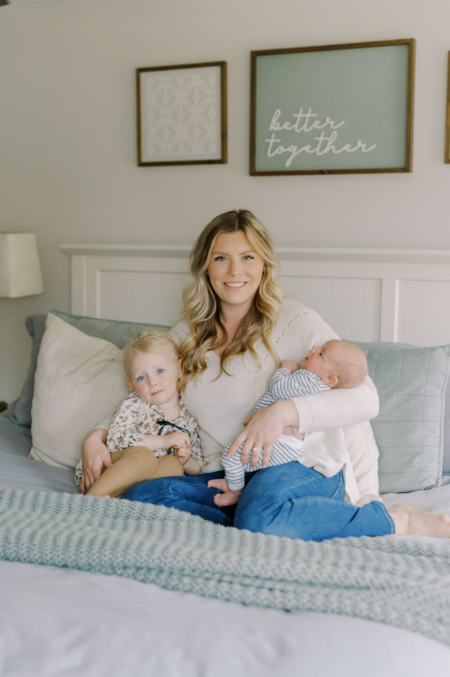 A smiling mother sits on a bed holding her two young children
