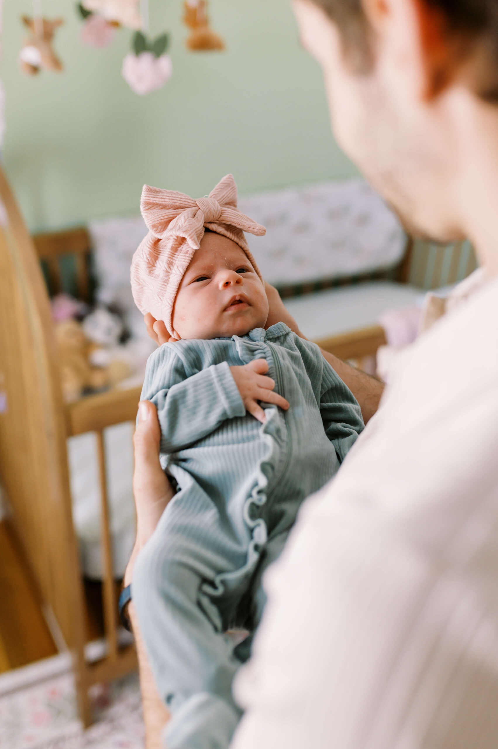 A look over dad's shoulder as he holds his newborn daughter in a blue onesie and pink bow in a nursery