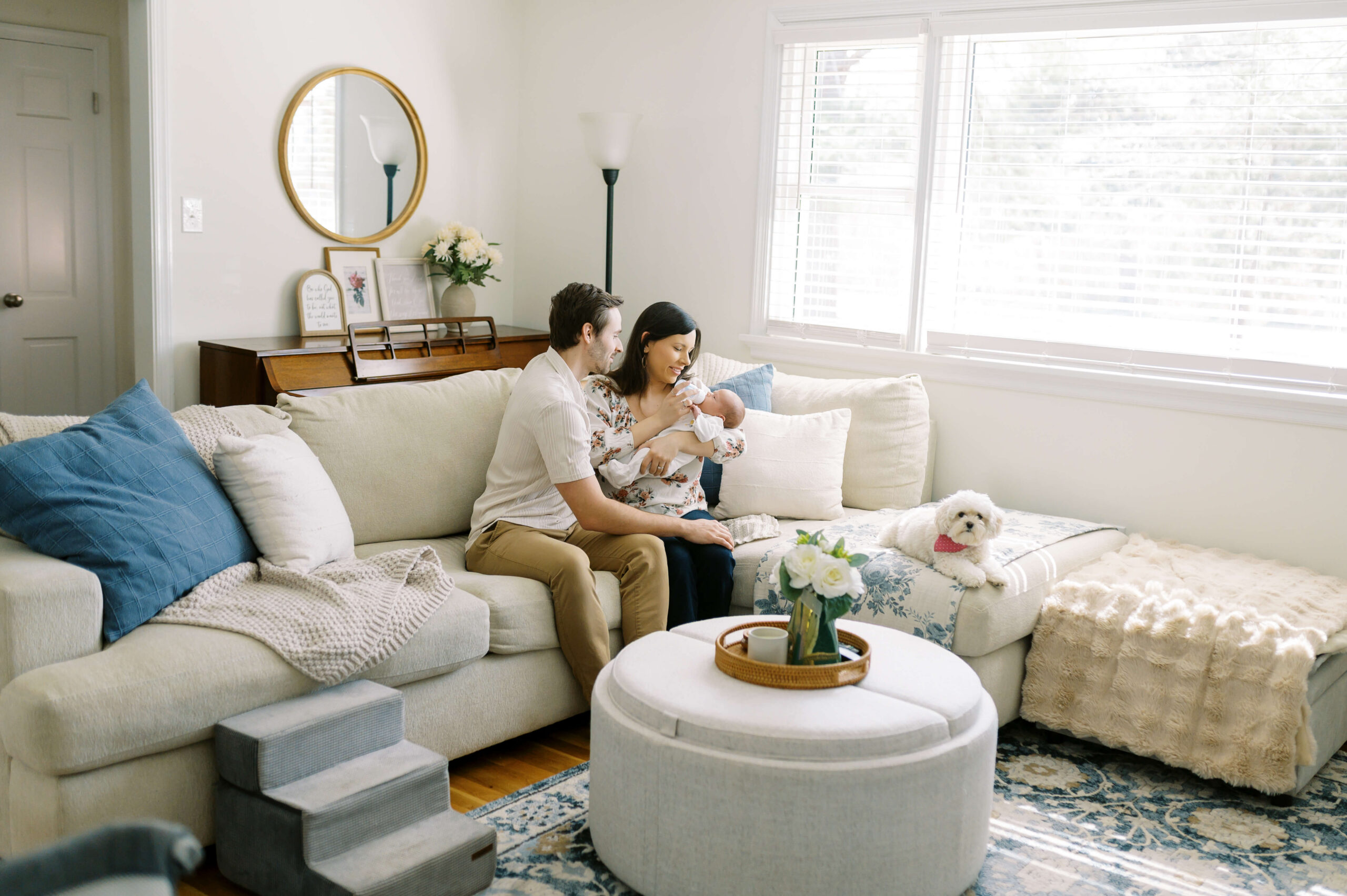 A mom and dad bottle feed their newborn on a couch with their small white dog