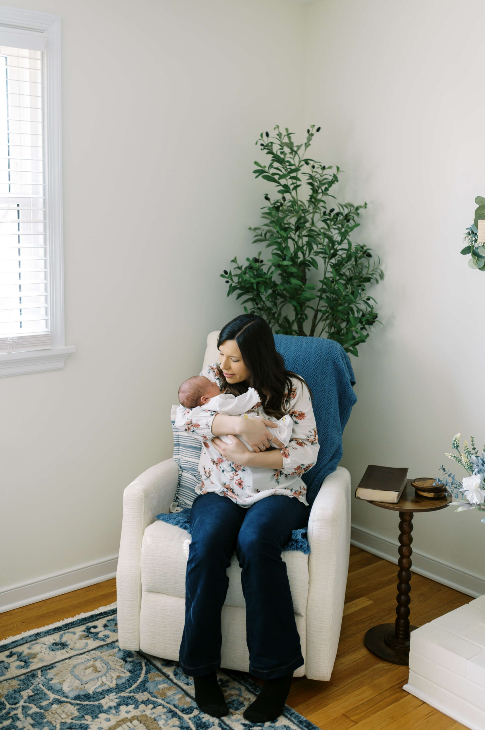 A happy new mom cradles her newborn in her arms while sitting in a nursing chair under a window thanks to postpartum doulas in Richmond, VA