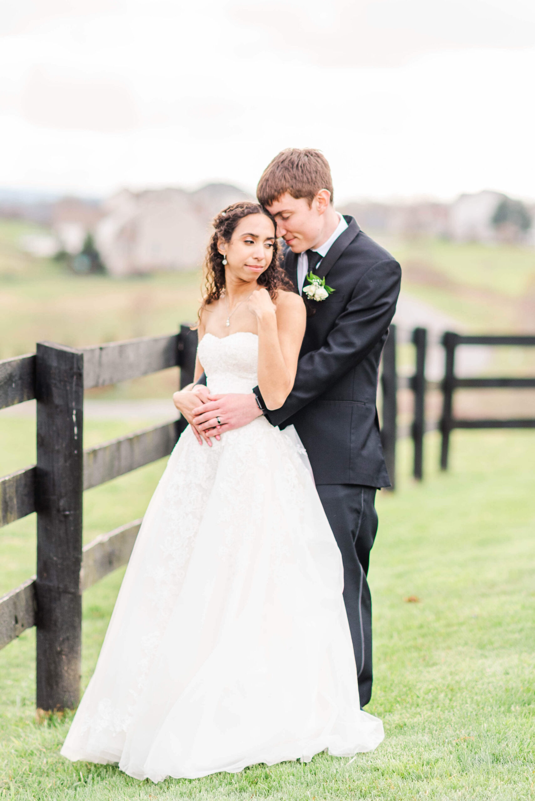 A bride is hugged from behind by her tall groom in black by a wooden fence during their shadow creek wedding