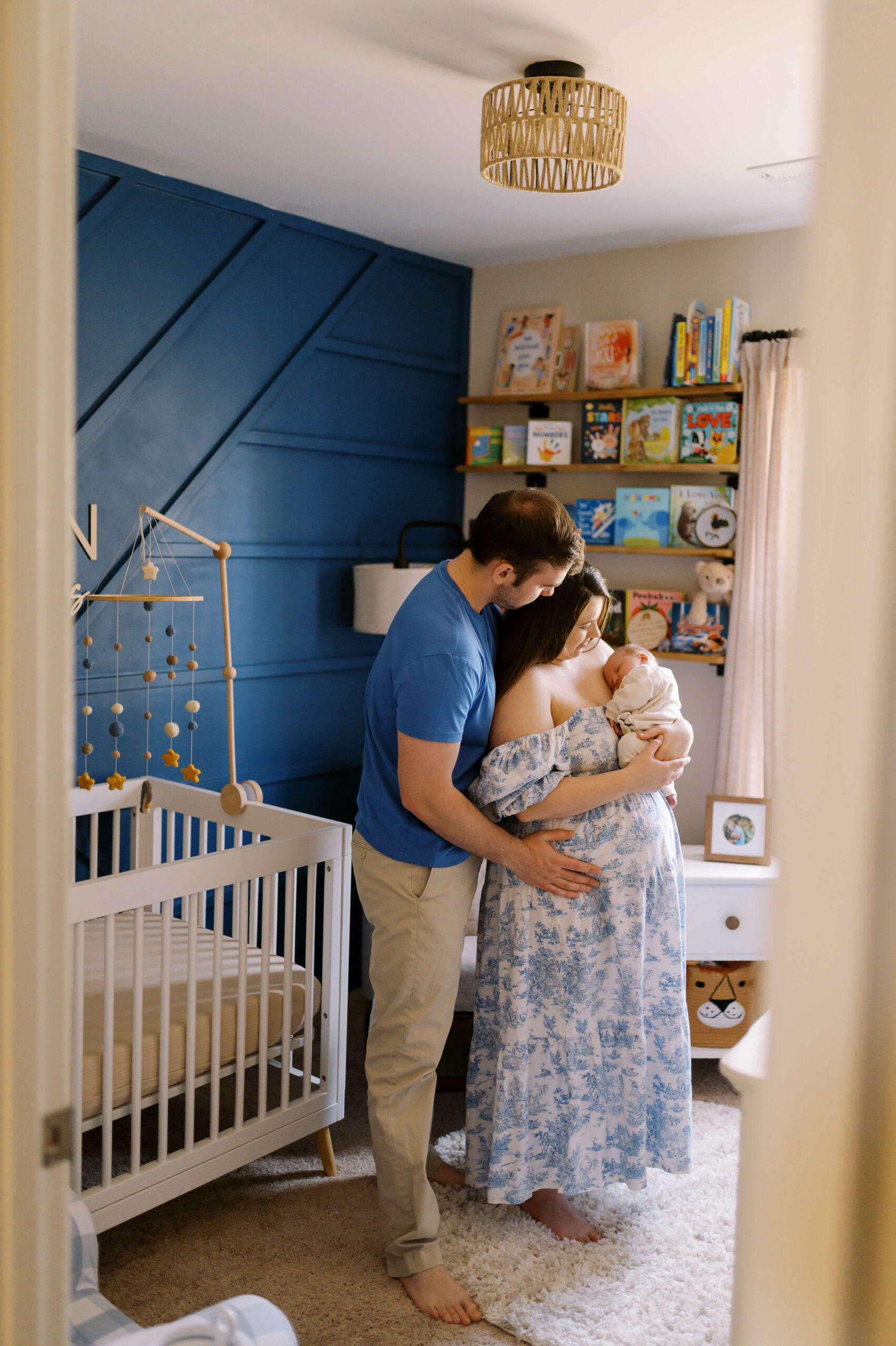 A happy mom and dad stand in a nursery holding their newborn sleeping on mom's shoulder in blue outfits thanks to sleep training in Richmond, VA