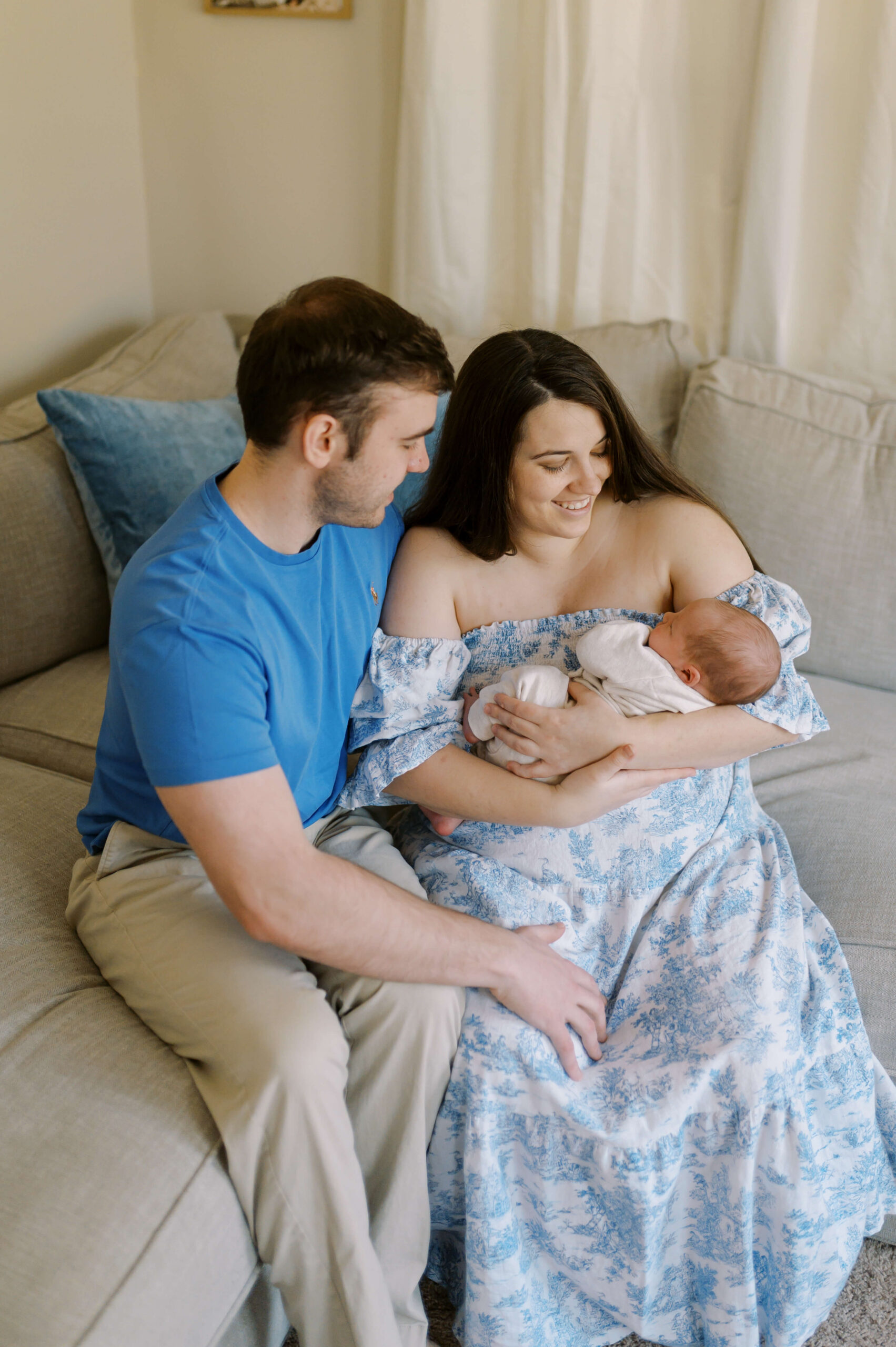 Happy new parents sit on a couch together in blue cradling their sleeping newborn baby in mom's arms