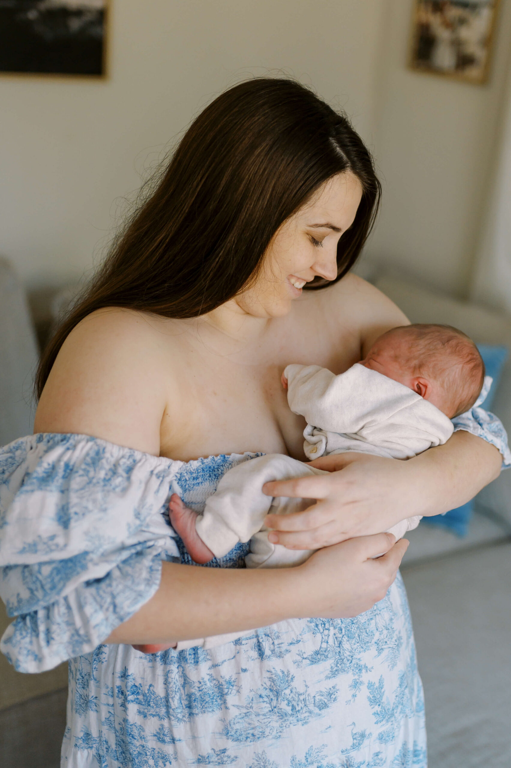 A happy new mom in a blue dress cradles her sleeping newborn while smiling in a window thanks to sleep training in Richmond, VA