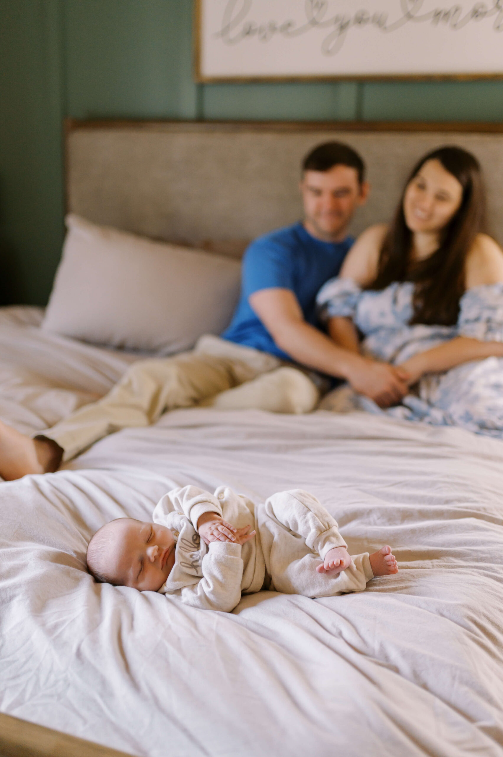 A newborn baby in a cream onesie sleeps on the corner of a bed as mom and dad sit behind watching thanks to sleep training in Richmond, VA