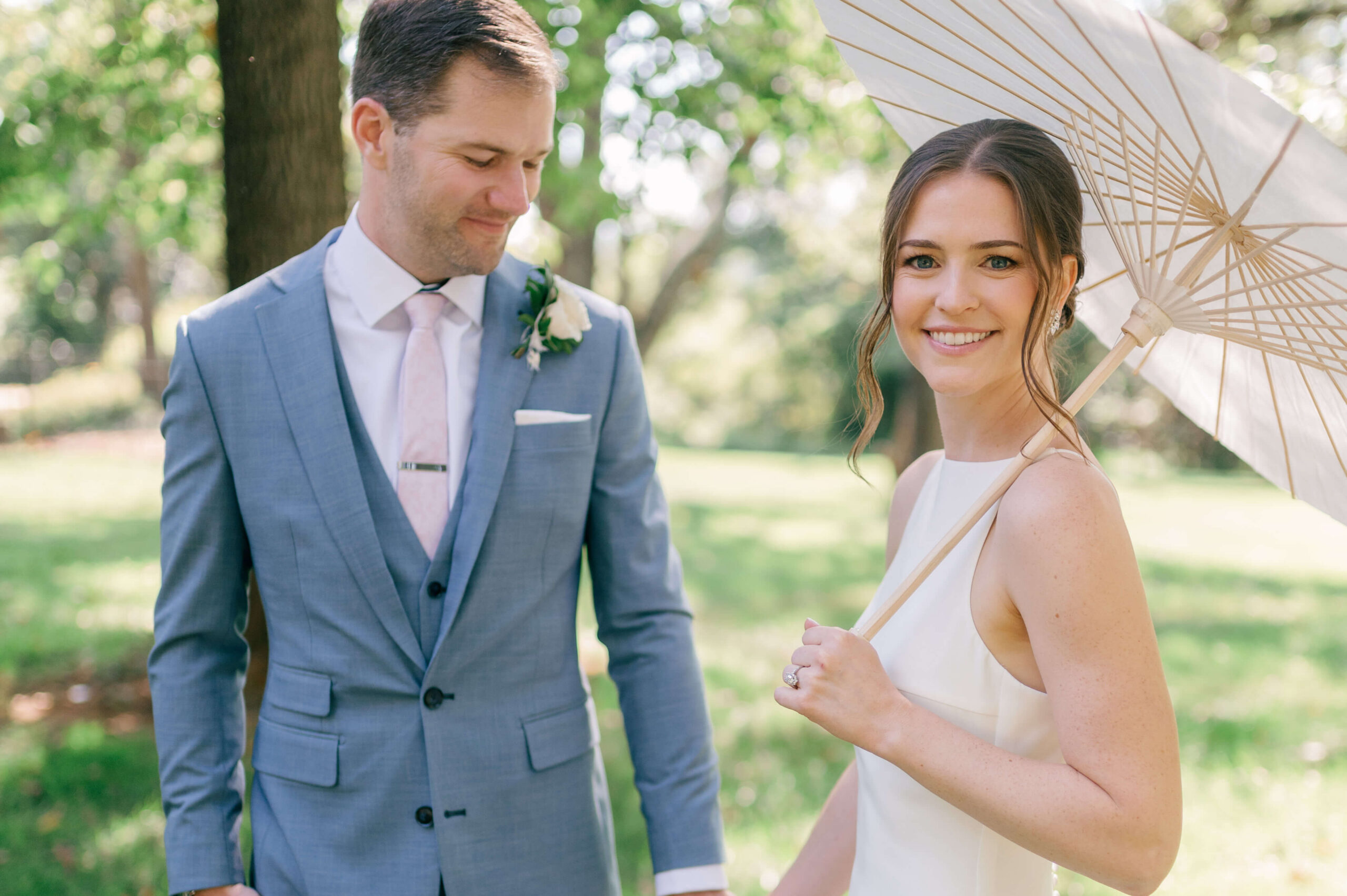 A bride walks in her elegant silk gown holding a parasol as her groom holds her hand