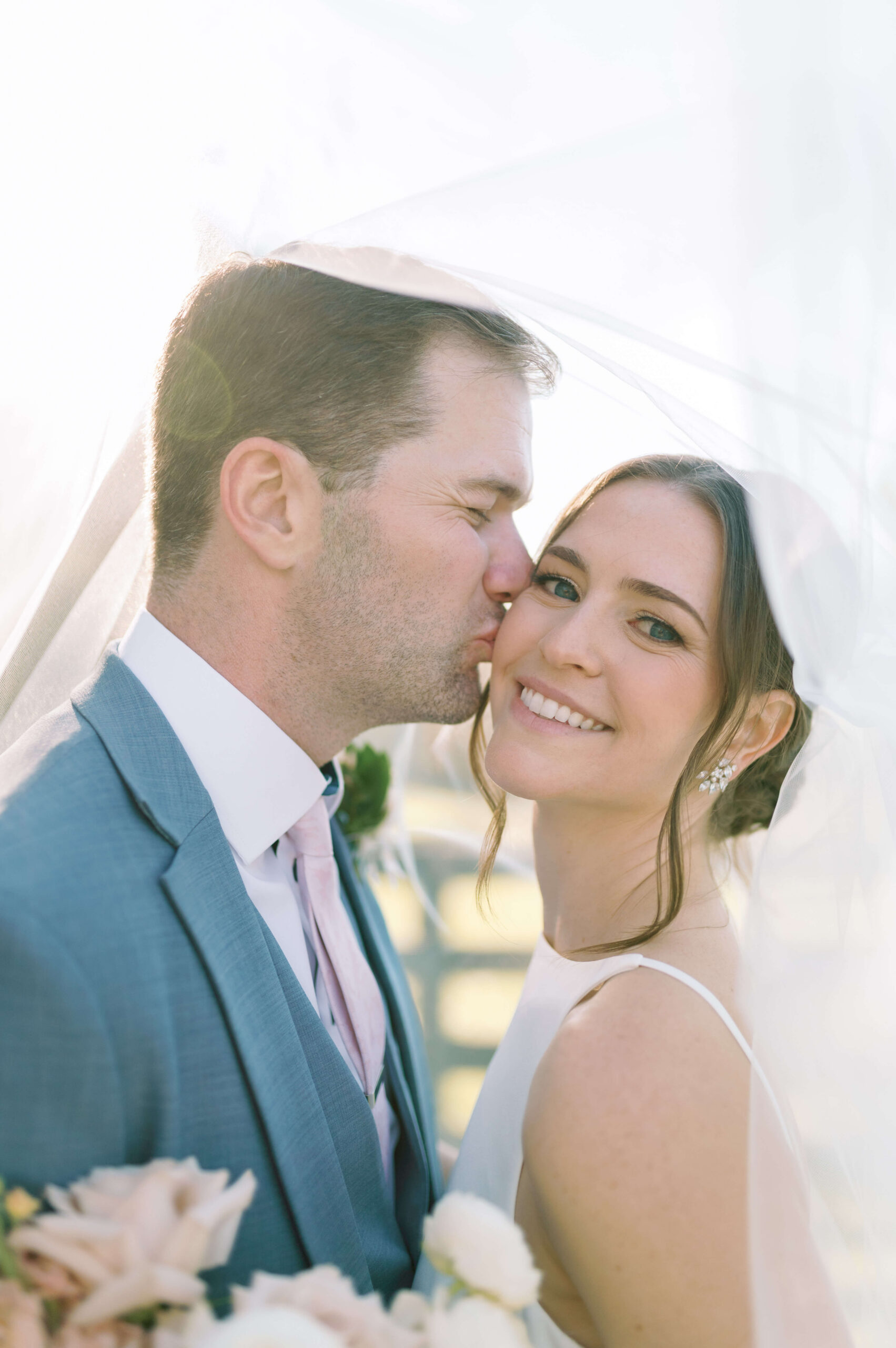 A groom in blue kisses the cheek of his happy bride under the veil at sunset during their veritas winery wedding